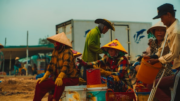 Several people wearing traditional conical hats are gathered in an outdoor setting, likely at a market or a coastal area. They appear to be engaged in sorting or handling seafood, with various baskets and containers around them. The individuals are dressed in colorful clothing, and a truck with a dolphin logo is visible in the background.
