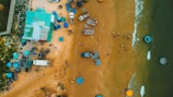 An aerial view of a coastal area where small vehicles, numerous blue round baskets, and other items are scattered on sandy terrain next to a body of water. The ocean is visible on one side with a few small circular boats floating nearby. Several people are dispersed around the sand, engaging in various activities.