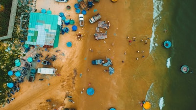 An aerial view of a coastal area where small vehicles, numerous blue round baskets, and other items are scattered on sandy terrain next to a body of water. The ocean is visible on one side with a few small circular boats floating nearby. Several people are dispersed around the sand, engaging in various activities.