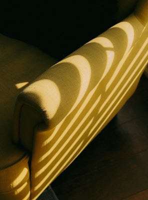 A close-up view of a mustard yellow armchair with sunlight streaming through blinds creating a pattern of light and shadow on the fabric. The shadows form curved stripes across the armrest and backrest.