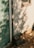 A teal green door with a modern keypad lock is set into a wooden outdoor wall. Dappled sunlight filters through leaves, casting soft shadows on the wall. The ground is covered with autumn leaves and a log is resting near the corner of the wall.