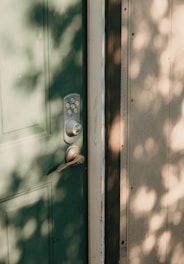 A green door with a modern keypad lockset and handle. Dappled sunlight creates soft shadows on the door and adjacent wall, enhancing the texture of the wood.