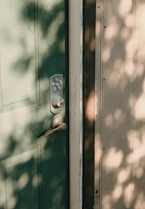 A green door with a modern keypad lockset and handle. Dappled sunlight creates soft shadows on the door and adjacent wall, enhancing the texture of the wood.
