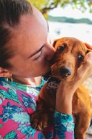 A person wearing a colorful floral shirt lovingly holds and kisses a small brown dog. The person gently cradles the dog’s head, showing a tender moment between them. The background features soft-focus greenery and a blurred body of water.