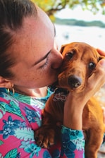 A person wearing a colorful floral shirt lovingly holds and kisses a small brown dog. The person gently cradles the dog’s head, showing a tender moment between them. The background features soft-focus greenery and a blurred body of water.
