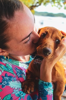 A person wearing a colorful floral shirt lovingly holds and kisses a small brown dog. The person gently cradles the dog’s head, showing a tender moment between them. The background features soft-focus greenery and a blurred body of water.