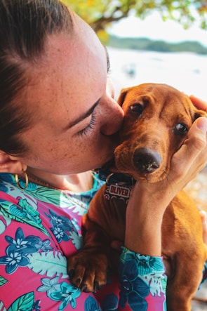 A person wearing a colorful floral shirt lovingly holds and kisses a small brown dog. The person gently cradles the dog’s head, showing a tender moment between them. The background features soft-focus greenery and a blurred body of water.