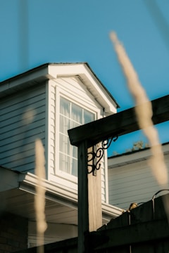 A close-up view of a house exterior showcasing a white, wood-paneled structure with a window on the attic. A wooden fence post with decorative metalwork is in the foreground, and tall grass is blurred in the foreground.