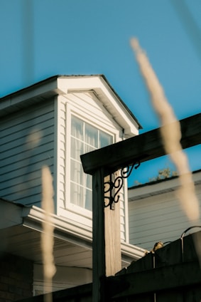 A close-up view of a house exterior showcasing a white, wood-paneled structure with a window on the attic. A wooden fence post with decorative metalwork is in the foreground, and tall grass is blurred in the foreground.