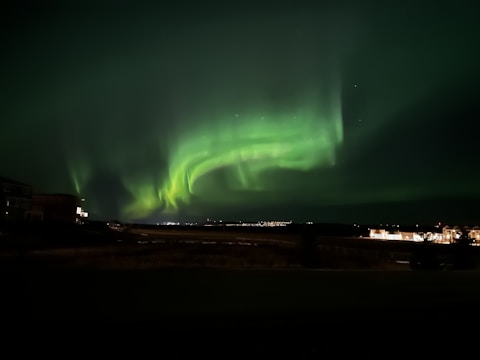 A mesmerizing display of green and yellow auroras sweeping across a dark night sky. The lights create swirling patterns over a landscape, which includes faint silhouettes of buildings in the foreground, illuminated by artificial lights.