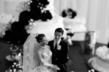 A black and white photo featuring a bride and groom cake topper in front of several cakes. The bride is wearing a detailed lace veil, and both figures are dressed in formal wedding attire. The background displays flowers and parts of wedding cakes, contributing to a celebration setting.