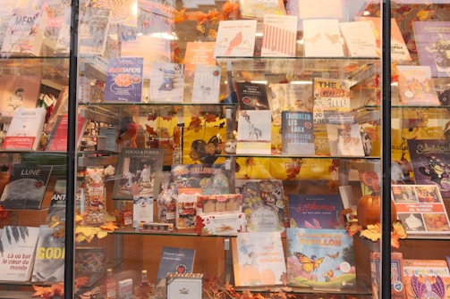 A bookstore display window featuring a variety of books and autumn-themed decorations. The shelves are filled with colorful book covers, including both fictional and non-fictional titles. Autumn leaves and small decorative pumpkins embellish the display, creating a cozy and inviting atmosphere. Some books are propped up while others lie flat, with a mix of vibrant and muted colors.