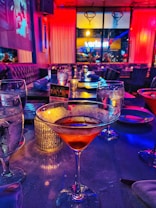 A dimly lit restaurant or bar setting featuring several tables with glassware. A cocktail in a martini glass is prominently in the foreground, while the background shows a vibrant mix of purple and red lighting. Large windows offer a view of the street outside, and a projected image is visible on one of the walls.
