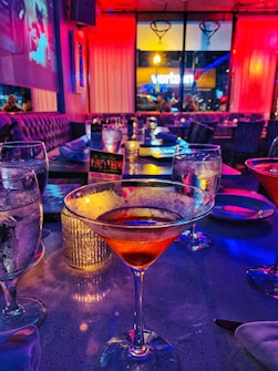 A dimly lit restaurant or bar setting featuring several tables with glassware. A cocktail in a martini glass is prominently in the foreground, while the background shows a vibrant mix of purple and red lighting. Large windows offer a view of the street outside, and a projected image is visible on one of the walls.
