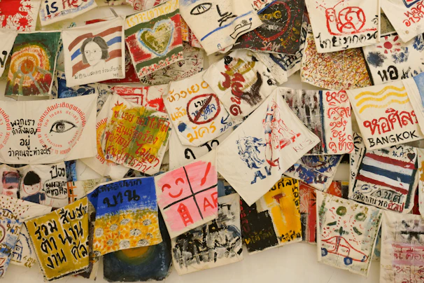 A collection of vibrant, hand-painted art pieces and illustrations on fabrics, featuring a mix of abstract designs, text, and depictions of various subjects. Some fabrics include written messages in different languages, alongside colorful imagery including hearts, eyes, faces, and various symbols.