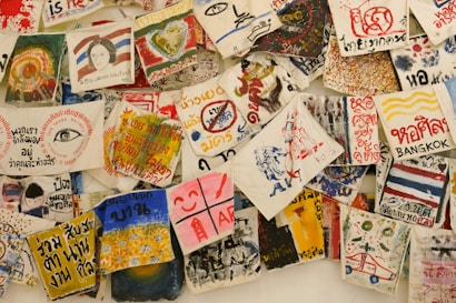 A collection of vibrant, hand-painted art pieces and illustrations on fabrics, featuring a mix of abstract designs, text, and depictions of various subjects. Some fabrics include written messages in different languages, alongside colorful imagery including hearts, eyes, faces, and various symbols.