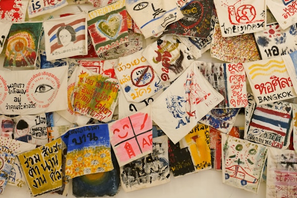 A collection of vibrant, hand-painted art pieces and illustrations on fabrics, featuring a mix of abstract designs, text, and depictions of various subjects. Some fabrics include written messages in different languages, alongside colorful imagery including hearts, eyes, faces, and various symbols.