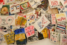 A collection of vibrant, hand-painted art pieces and illustrations on fabrics, featuring a mix of abstract designs, text, and depictions of various subjects. Some fabrics include written messages in different languages, alongside colorful imagery including hearts, eyes, faces, and various symbols.