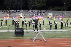 A marching band performs on a football field at night, with band members playing brass instruments and waving colorful flags. A conductor stands on a ladder, leading the ensemble. In the background, a crowd sits in bleachers, watching the performance. Painted backdrops depicting clouds and sky are also visible.