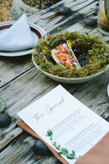 A rustic wooden table displays a plate garnished with moss, holding a colorful taco with various vegetables and garnishes. A neatly folded napkin is placed on another plate beside it. A menu titled 'The Special' is laid on the table with two dish options, accompanied by green leaves as decoration. River stones and a glass of a layered beverage can also be seen in this outdoor dining setting.