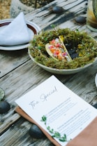A rustic wooden table displays a plate garnished with moss, holding a colorful taco with various vegetables and garnishes. A neatly folded napkin is placed on another plate beside it. A menu titled 'The Special' is laid on the table with two dish options, accompanied by green leaves as decoration. River stones and a glass of a layered beverage can also be seen in this outdoor dining setting.