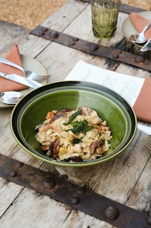 A rustic setting with a wooden table, featuring a green bowl filled with pasta garnished with herbs and mushrooms. Nearby are plates with folded napkins and cutlery, as well as a green glass and a menu.