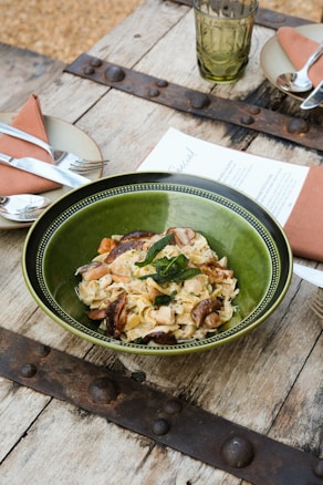 A rustic setting with a wooden table, featuring a green bowl filled with pasta garnished with herbs and mushrooms. Nearby are plates with folded napkins and cutlery, as well as a green glass and a menu.