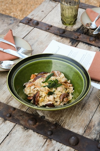 A rustic setting with a wooden table, featuring a green bowl filled with pasta garnished with herbs and mushrooms. Nearby are plates with folded napkins and cutlery, as well as a green glass and a menu.