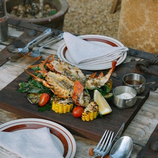 A wooden board serves grilled lobster halves garnished with herbs, surrounded by charred corn, cherry tomatoes, and a lemon wedge. Metal cups with sauces accompany the dish. The rustic wooden table is set with plates, napkins, and cutlery, creating an inviting outdoor dining scene.