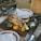 A wooden board serves grilled lobster halves garnished with herbs, surrounded by charred corn, cherry tomatoes, and a lemon wedge. Metal cups with sauces accompany the dish. The rustic wooden table is set with plates, napkins, and cutlery, creating an inviting outdoor dining scene.