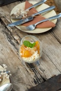 A rustic wooden table holds a dessert presentation featuring a glass filled with creamy dessert topped with mango pieces and a sprig of mint. Nearby, a small plate with a folded napkin, cutlery, and delicate baby's breath flowers adds a touch of elegance.