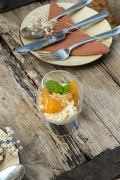 A rustic wooden table holds a dessert presentation featuring a glass filled with creamy dessert topped with mango pieces and a sprig of mint. Nearby, a small plate with a folded napkin, cutlery, and delicate baby's breath flowers adds a touch of elegance.