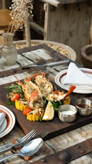 A wooden table is set with a rustic and gourmet meal consisting of grilled lobster, corn on the cob, vegetables, and a lemon wedge placed on a wooden serving board. Metal utensils, a stack of plates with a folded napkin, and small containers of sauces are also on the table. A glass vase with dried white flowers is part of the table decor. The background contains wooden elements, suggesting an outdoor dining setting.