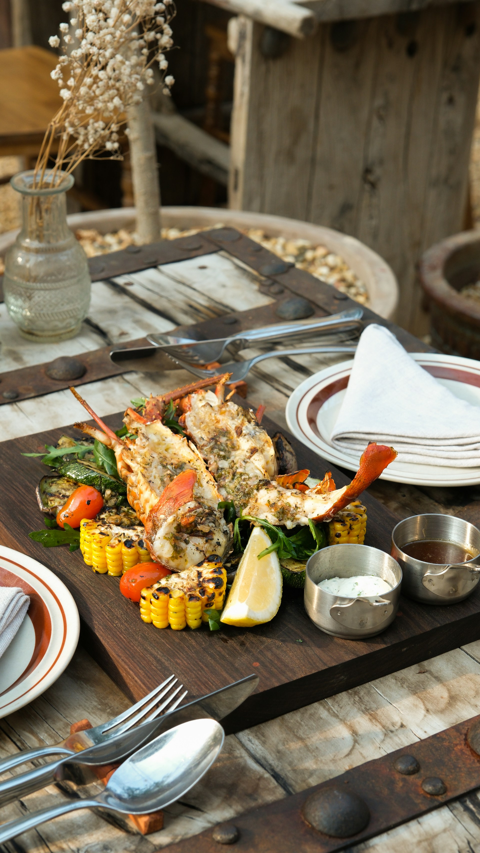 A wooden table is set with a rustic and gourmet meal consisting of grilled lobster, corn on the cob, vegetables, and a lemon wedge placed on a wooden serving board. Metal utensils, a stack of plates with a folded napkin, and small containers of sauces are also on the table. A glass vase with dried white flowers is part of the table decor. The background contains wooden elements, suggesting an outdoor dining setting.