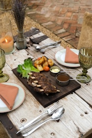 An elegantly set outdoor dining table featuring a wooden board with grilled ribs, roasted potatoes, lettuce leaves, and dipping sauces. There are two plates with salmon-colored napkins, green-tinted glasses, silver cutlery, and a lit candle in a glass holder. The background includes a rustic brick floor and a vase with dried flowers.