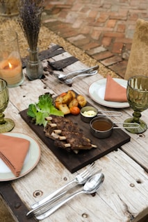 An elegantly set outdoor dining table featuring a wooden board with grilled ribs, roasted potatoes, lettuce leaves, and dipping sauces. There are two plates with salmon-colored napkins, green-tinted glasses, silver cutlery, and a lit candle in a glass holder. The background includes a rustic brick floor and a vase with dried flowers.