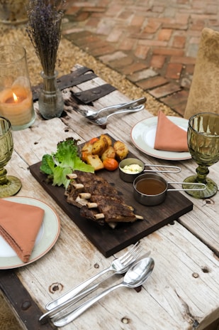 An elegantly set outdoor dining table featuring a wooden board with grilled ribs, roasted potatoes, lettuce leaves, and dipping sauces. There are two plates with salmon-colored napkins, green-tinted glasses, silver cutlery, and a lit candle in a glass holder. The background includes a rustic brick floor and a vase with dried flowers.