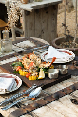 A rustic wooden butcher block table displaying an inviting spread of Acadian dishes including seafood platter, smoked meat, and blueberry tart, styled with mid-century modern tableware.
