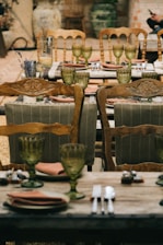 An elegantly set outdoor dining table featuring vintage wooden chairs and green tinted glass goblets. The table is arranged with earth-toned plates, cloth napkins, and polished cutlery, creating a rustic yet sophisticated ambiance. Decor includes dried lavender and candles, complementing the brick and natural elements in the background.