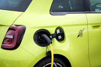 The image features the rear section of a bright yellow electric vehicle with a charging plug inserted into the charge port. The taillight design is prominent, and there is a distinctive logo near the charge port.