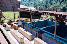 A rustic outdoor washing station with multiple tiled basins, shaded by a corrugated metal roof. The setup includes several pipes and exposed wooden structures with signs labeling different sections. The background shows a grassy area, a fence made of wooden poles, and a brick building under a clear sky.