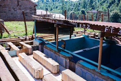 A rustic outdoor washing station with multiple tiled basins, shaded by a corrugated metal roof. The setup includes several pipes and exposed wooden structures with signs labeling different sections. The background shows a grassy area, a fence made of wooden poles, and a brick building under a clear sky.