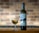 A tall bottle of Assyrtiko wine is placed next to a filled wine glass. The label on the bottle features modern, abstract artwork with a blue circle and black shapes. Both are set against a brick wall background, which is slightly out of focus.