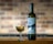 A tall bottle of Assyrtiko wine is placed next to a filled wine glass. The label on the bottle features modern, abstract artwork with a blue circle and black shapes. Both are set against a brick wall background, which is slightly out of focus.