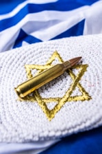 A brass bullet rests on a white crocheted fabric, which features a gold embroidered Star of David. The background displays a blue and white flag.