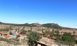A picturesque rural landscape featuring a small village with red-roofed houses surrounded by trees and fields, against a backdrop of rolling hills and a clear, blue sky.