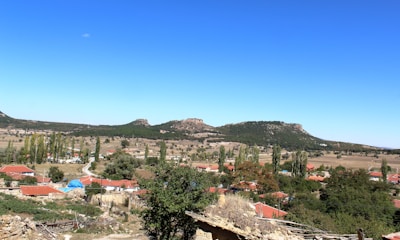 A picturesque rural landscape featuring a small village with red-roofed houses surrounded by trees and fields, against a backdrop of rolling hills and a clear, blue sky.