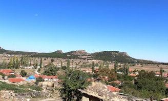 A picturesque rural landscape featuring a small village with red-roofed houses surrounded by trees and fields, against a backdrop of rolling hills and a clear, blue sky.