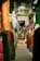 A narrow aisle in a bustling clothing market, with a variety of colorful garments hanging on both sides. The market appears crowded, with racks full of dresses, shirts, and traditional garments. A sign above reads 'Song Jaya Fashion.' The setting suggests a lively, busy environment typical of traditional markets.
