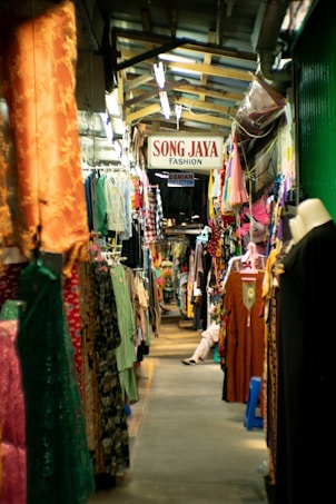 A narrow aisle in a bustling clothing market, with a variety of colorful garments hanging on both sides. The market appears crowded, with racks full of dresses, shirts, and traditional garments. A sign above reads 'Song Jaya Fashion.' The setting suggests a lively, busy environment typical of traditional markets.